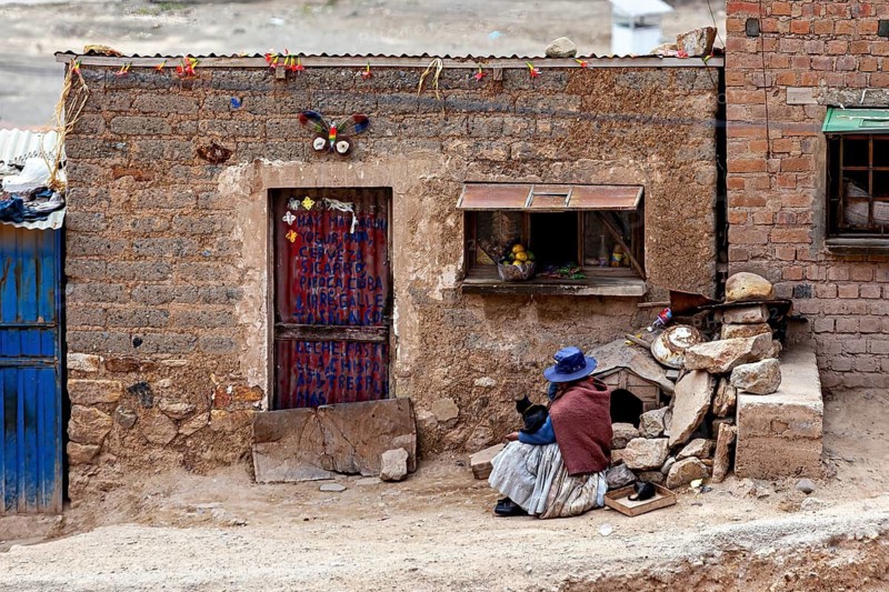 A woman in traditional dress sits on a floor in front of a brick wall with a decorated door, a woman at her house by the silver mines near Potosi on Cerro Rico in Bolivia