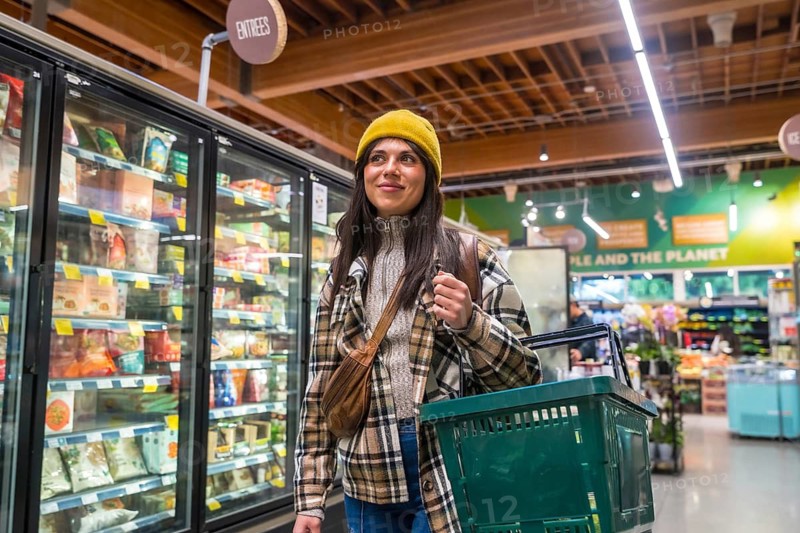 Woman in a yellow beanie and plaid shirt walking through a modern grocery store aisle, carrying a shopping basket and looking at frozen food options, representing daily life and consumerism