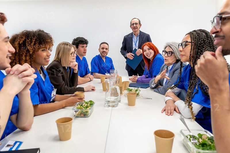 Diverse group of medical professionals, including doctors and nurses, collaborating and discussing ideas during an informal meeting with healthy lunch in a bright clinic setting