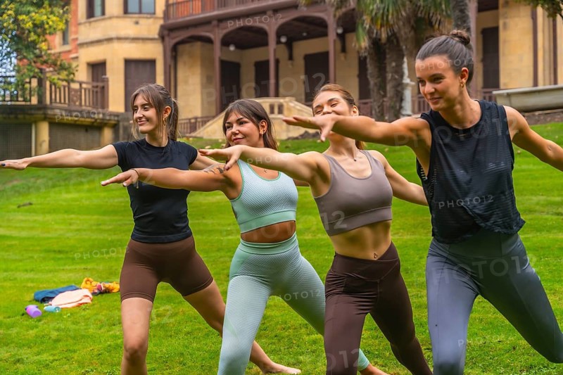Young women practicing yoga in a park, balancing in warrior poses and stretching together on green grass, enjoying mindfulness, fitness, and friendly outdoor wellness