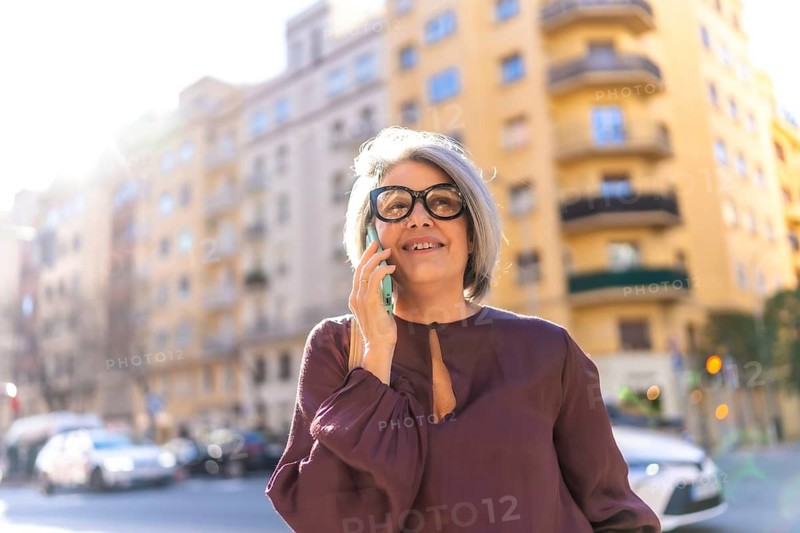 Smiling mature woman with glasses and gray hair talking on a mobile phone, representing modern communication and an active urban senior lifestyle in a bright city street