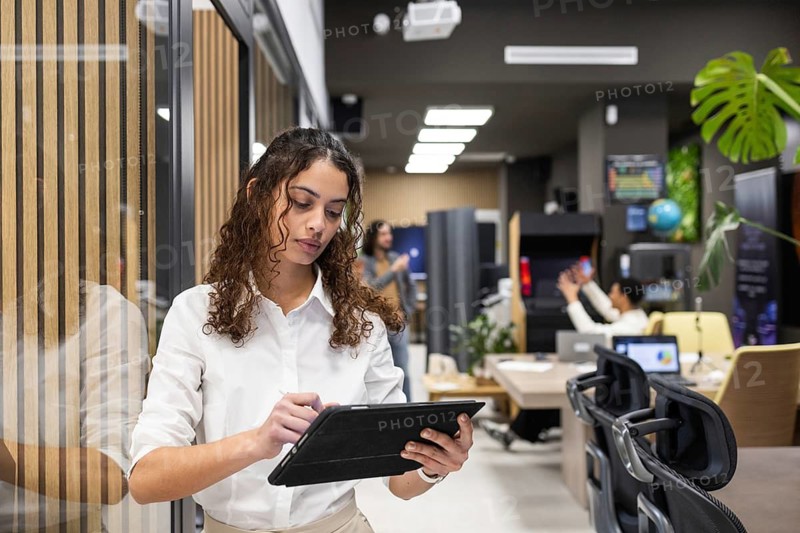 A professional woman in a white shirt uses a tablet in a stylish, modern office. Colleagues work collaboratively in the background, showcasing a tech-driven work environment