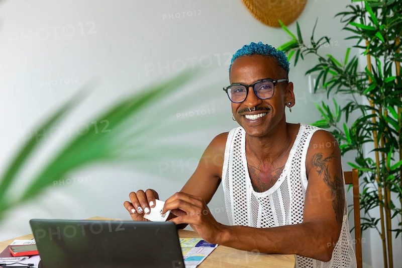 A joyful LGBTQIA+ individual with blue hair and glasses is preparing for holidays at home, surrounded by greenery and working on their laptop