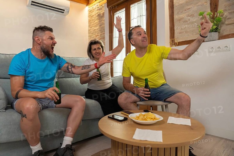 Three friends react passionately while watching a sports game at home, cheering and arguing with beers and chips on the table in a lively living room scene