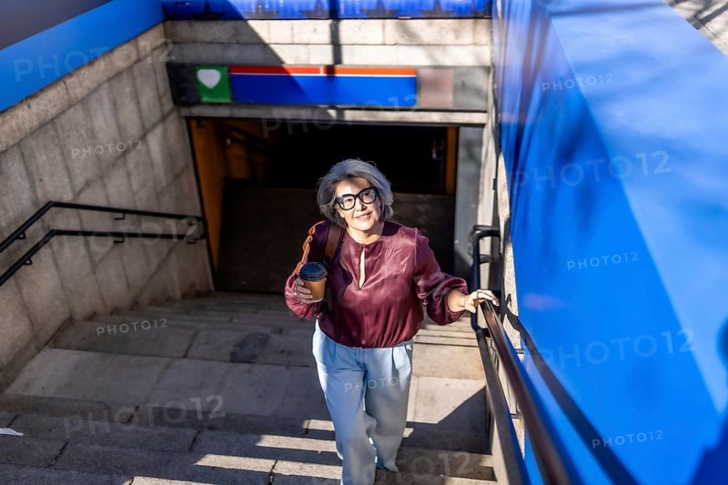 Senior woman commuters navigating subway stairs, holding a disposable coffee cup and backpack, embodying urban lifestyle and modern senior engagement in city travel