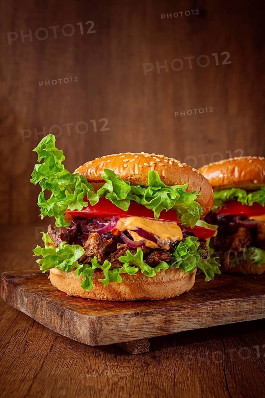 Two burgers are stacked high with fresh lettuce, tomato slices, grilled onions, and a savory sauce. They sit on a wooden board in a warm light setting. Ideal for a meal or snack
