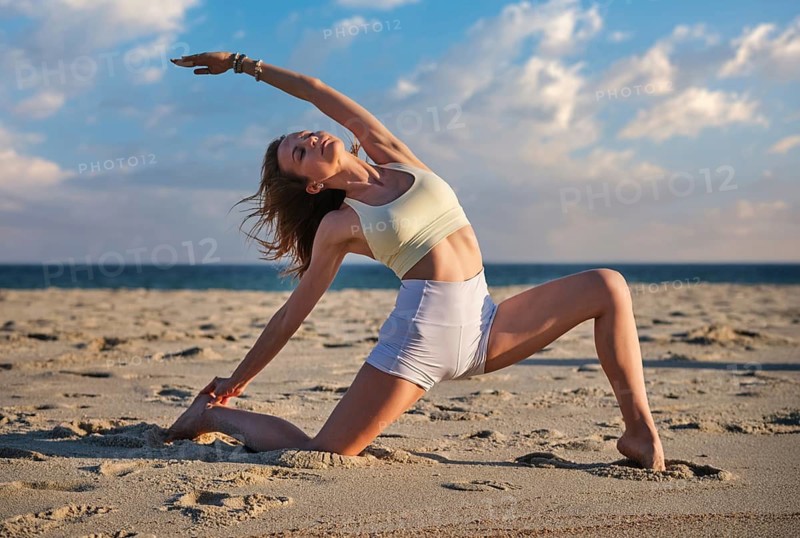 Female yoga practitioner in low crescent lunge at the shoreline, stretching deeply on sunlit sand with ocean horizon and dramatic clouds behind