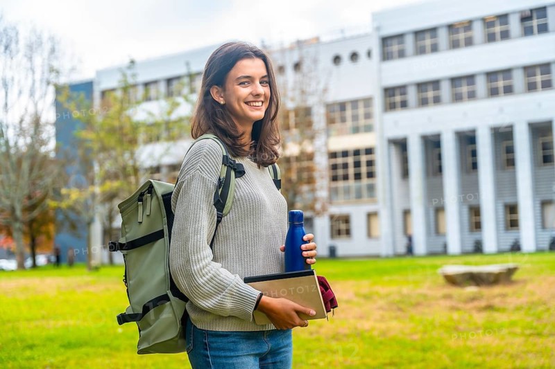 Female student carrying a backpack and books while holding a water bottle, standing on a green lawn with a modern university building in the background, representing education and academic life