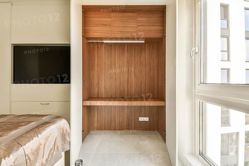 A modern bedroom featuring a luxurious wooden wardrobe with ample shelves, a wall-mounted TV, and a plush bedspread. The room is warmly lit by natural sunlight through a large window, creating a cozy atmosphere