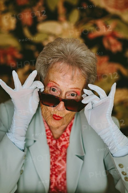 A lively elderly woman strikes a playful pose with sunglasses in a studio. Her vibrant outfit, gloves, and lipstick showcase her bold, youthful spirit, set against a floral backdrop