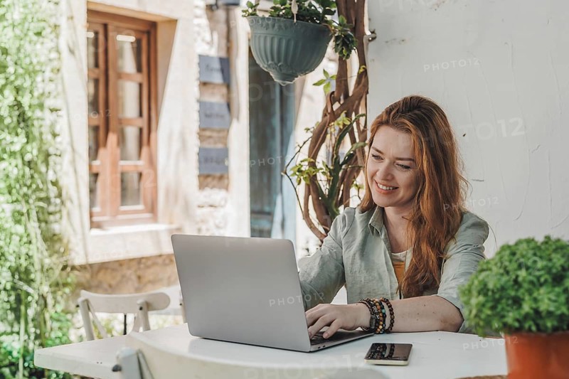 A woman working on a laptop at an outdoor table, surrounded by greenery. She is focused and smiling, enjoying remote work in a relaxed setting