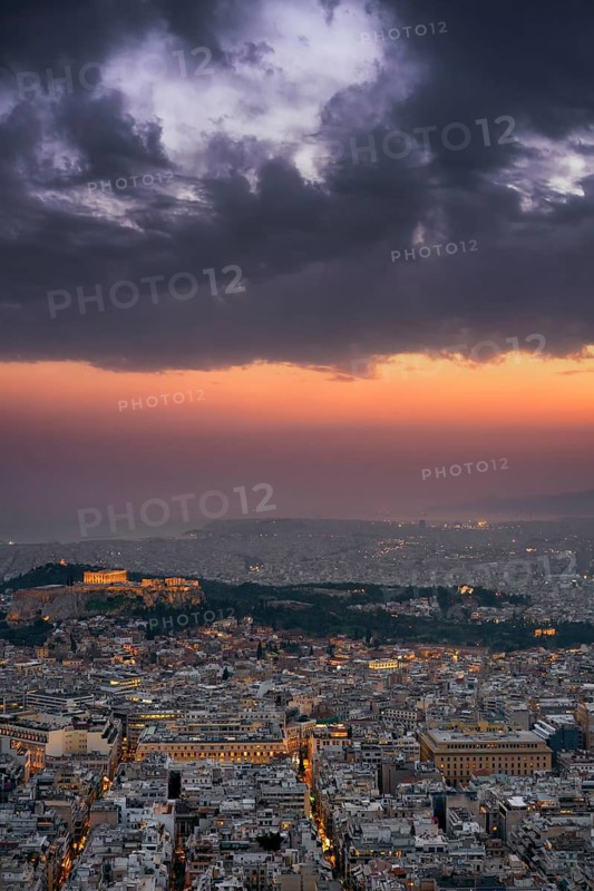 A dramatic sunset over Athens, Greece, with the Parthenon and Acropolis illuminated on the hilltop. The city lights gradually awaken as dark clouds fill the colorful sky, creating a striking urban scene