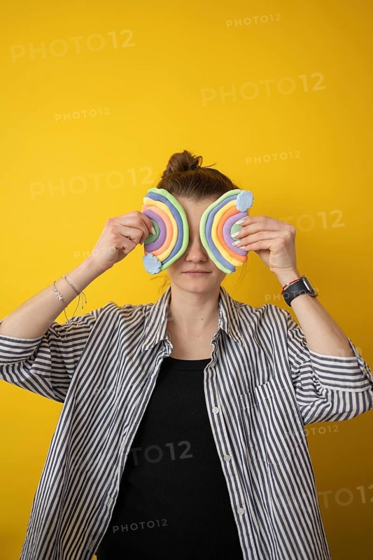 A person holds colorful clay rainbow shapes over their eyes, standing before a bright yellow backdrop. The playful expression embodies creativity and imagination