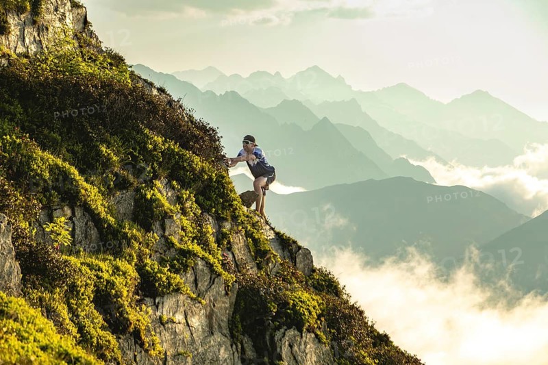 Side view of a trail runner scales a steep, rocky incline on a sunlit mountain. Surrounded by lush greenery, the runner wears athletic gear, sunglasses, and a visor, embodying adventure