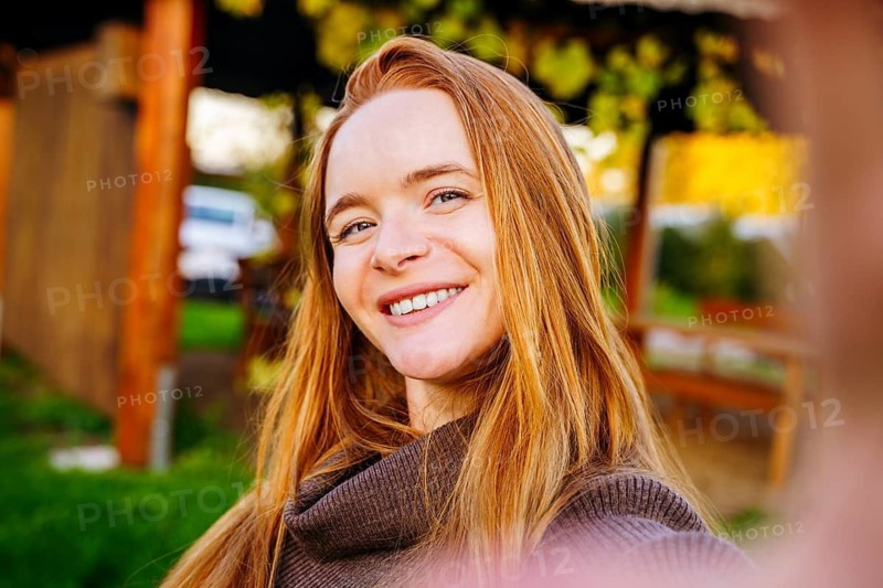 A smiling redhead woman takes a cheerful selfie outdoors, surrounded by lush greenery and warm sunlight. Her long hair and cozy sweater add to the relaxed, joyful scene