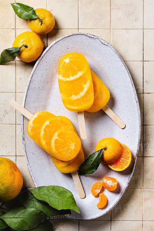 Top view of a refreshing tangerine flavored ice pops displayed on a rustic ceramic plate, complemented by fresh tangerines and green leaves
