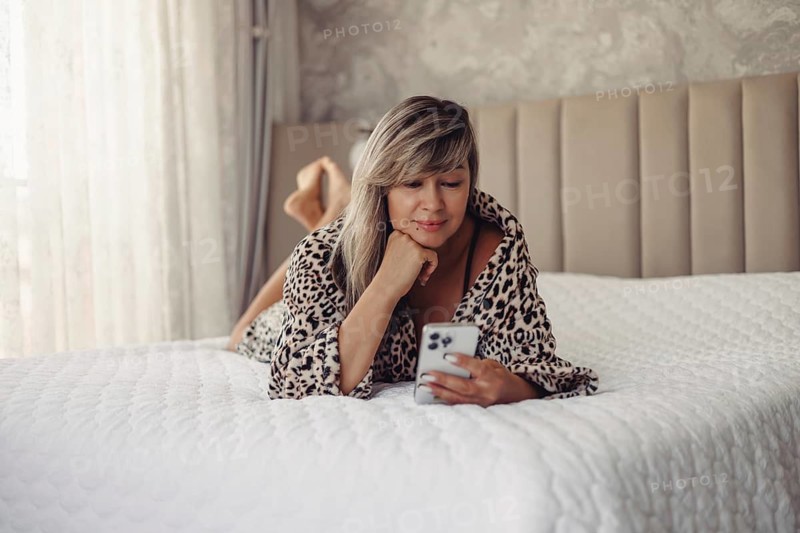 A woman in a leopard print robe lies on a white bed while using her smartphone. She looks content and relaxed, surrounded by a comfortable and cozy bedroom setting