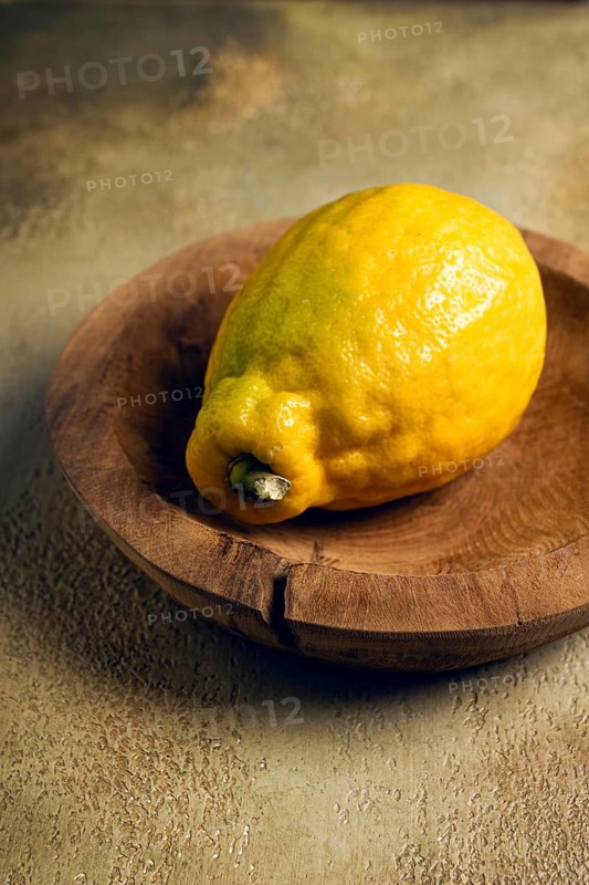 A vibrant yellow lemon sits in a hand-carved wooden bowl. The lemon\'s textured skin shines in natural light, highlighting its freshness and color against the rustic backdrop