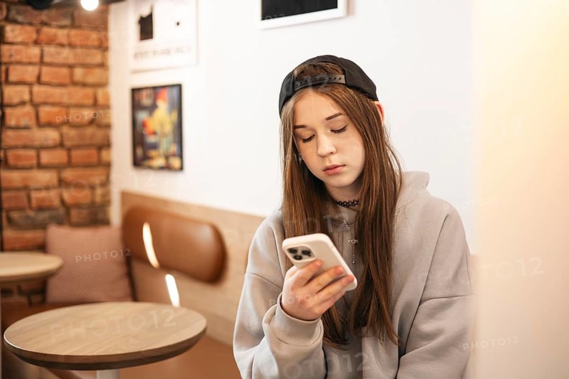 Teenage girl wearing casual clothing, intently focused on her smartphone while seated in a cozy cafe. The setting is relaxed with warm brick walls and soft lighting