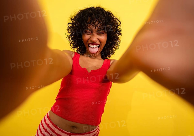Playful young woman taking a selfie against a bright yellow background, wearing a red top and striped shorts. She sticks out her tongue, creating a fun and energetic vibe