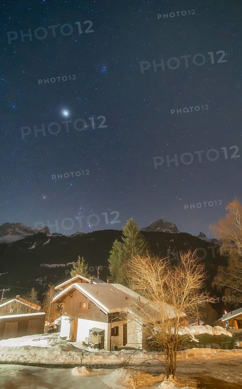 A starry night sky showcases the constellations of Orion and Taurus, along with Jupiter, the Pleiades cluster, and the bright star Sirius. A cozy snow-covered cabin adds warmth to the alpine scene