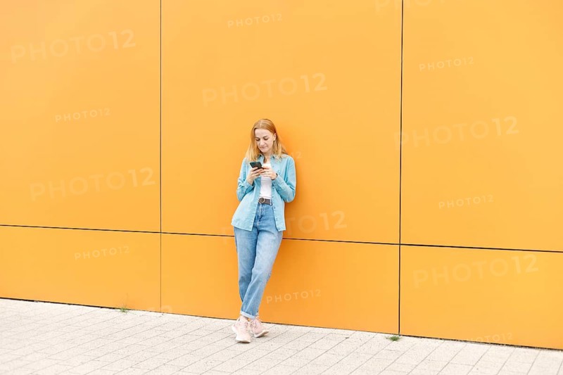 A woman in casual attire leans against a vibrant yellow wall, engaging with her smartphone The scene captures a modern urban vibe in Warsaw, blending technology with style
