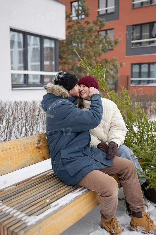A couple sits on a snow-dusted bench outside an apartment building, bundled in winter coats and hats, sharing a joyful moment The evergreen fir add to the festive atmosphere