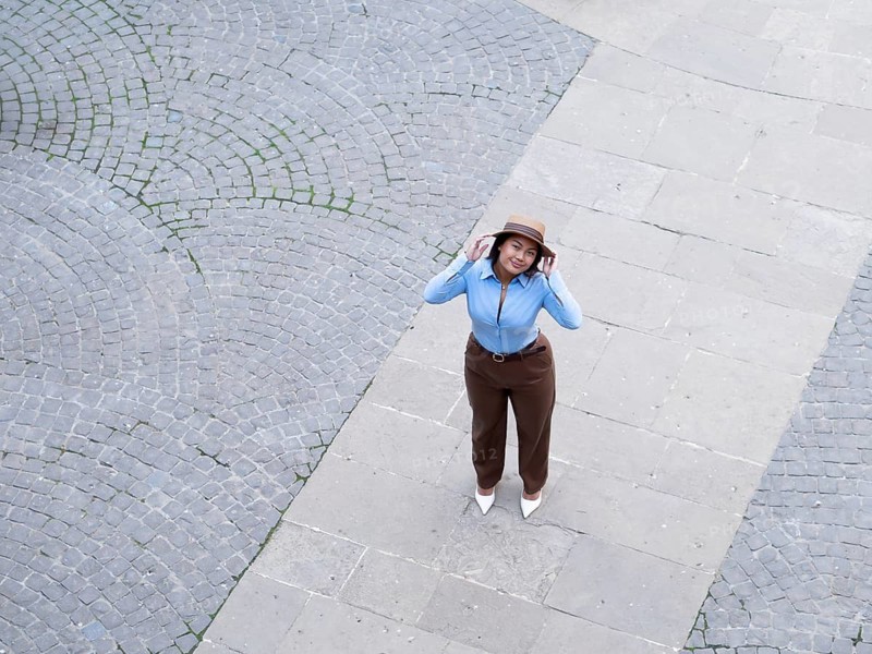 A high-angle view of a woman dressed in a blue shirt and brown pants, adjusting her hat on a cobblestone street, looking directly at the camera with a smile
