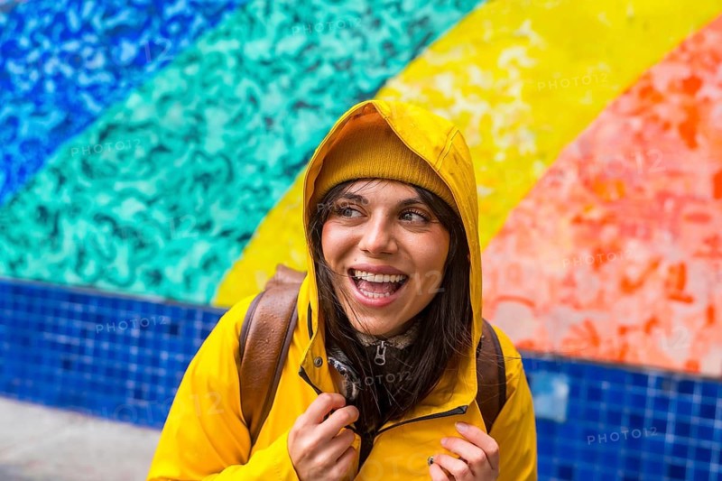 Happy young woman wearing a yellow raincoat and matching beanie, laughing and looking away, standing in front of a vibrant rainbow mural in urban san francisco