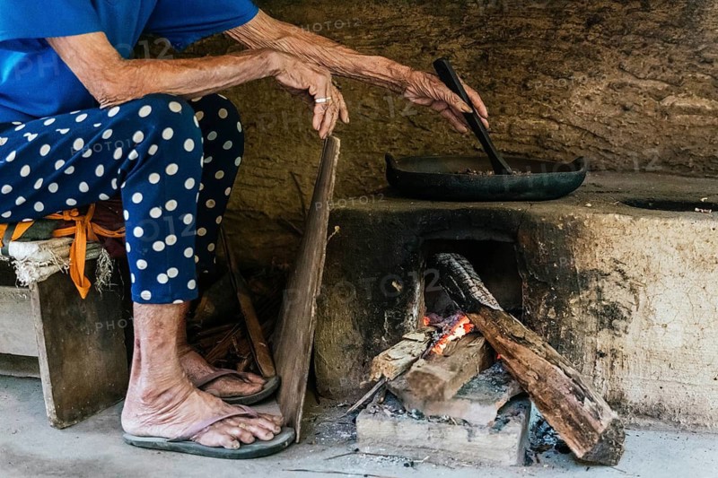 An elderly woman cooks balinese coffee over a wood fired stove in a rustic setting. The scene evokes a sense of tradition and simplicity, highlighting age old cooking techniques