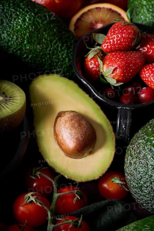 A vibrant display of fresh produce featuring ripe avocados, strawberries, kiwis, tomatoes, and blood oranges. The assortment highlights a mix of colors and textures