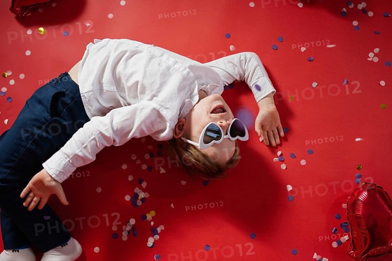 A young male child lies on a bright red background, joyfully wearing oversized heart-shaped sunglasses. Valentine's themed confetti and balloons enhance the festive mood