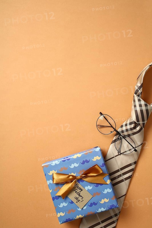 Top view of a colorful gift box with a golden bow and Happy Father's Day tag, accompanied by a stylish checkered tie and round glasses, all arranged on a warm orange backdrop. Perfect for celebrating dad