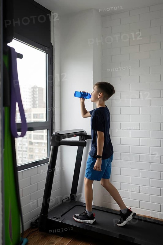 A boy in blue shorts and a dark shirt drinks water while on a treadmill next to a bright window. He stays hydrated and focused, showcasing a healthy exercise routine at home