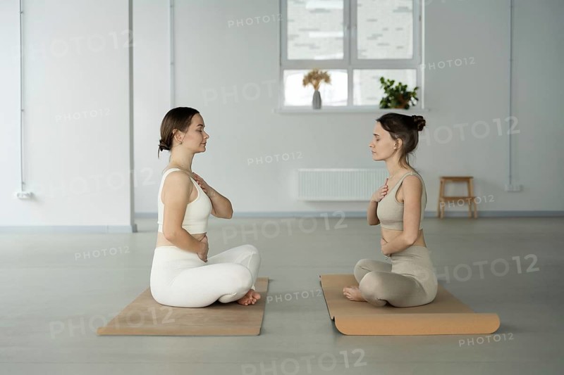 Two women sit cross legged on yoga mats in a serene yoga studio, engaging in mindfulness meditation with hands on their chests, promoting calmness and relaxation