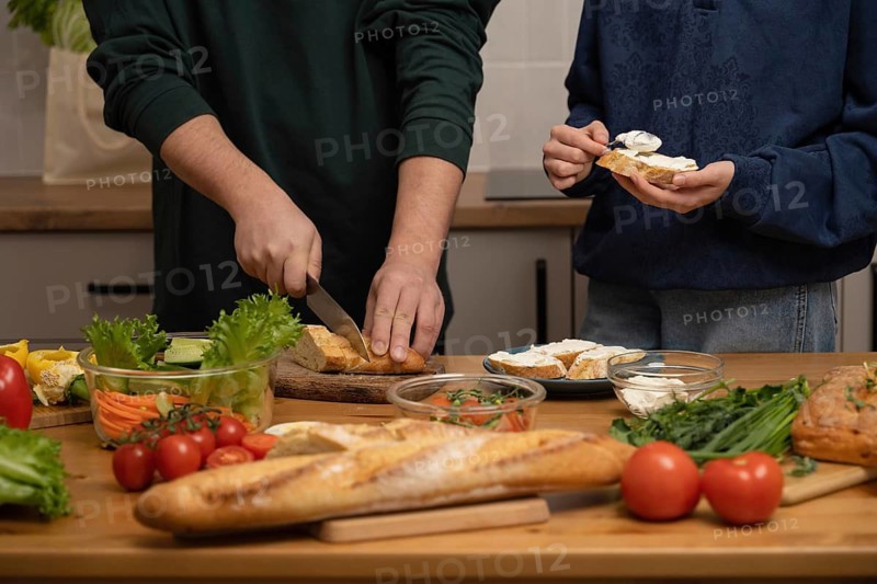 People preparing a fresh sandwich with a crusty baguette, cream cheese, and various vegetables. Vibrant ingredients and hands in action create a warm, inviting kitchen scene
