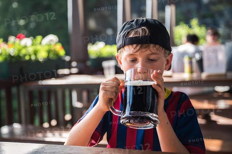 Cute boy is drinking a black drink from a large mug at a restaurant outdoor table