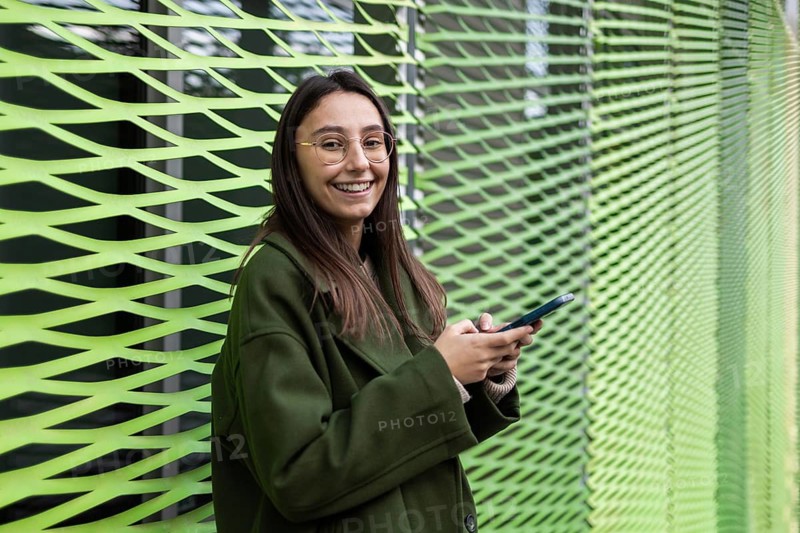 A woman wearing a green coat smiles as she uses her smartphone outside The lively green backdrop adds vibrancy, creating a cheerful and modern urban setting