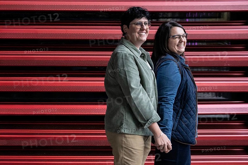 A joyful, curvy lesbian couple holding hands and smiling, standing in front of a modern red patterned wall. They express love, diversity, and unity in an urban setting