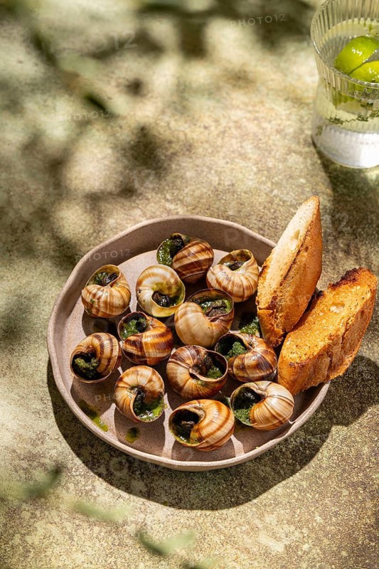 A gourmet plate of baked escargot generously topped with melted butter and fresh parsley, accompanied by crisp, toasted bread slices