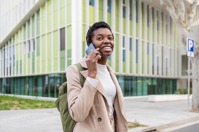 A stylish black woman confidently talks on her phone while walking in an urban environment She wears a beige coat and smiles, exuding positivity