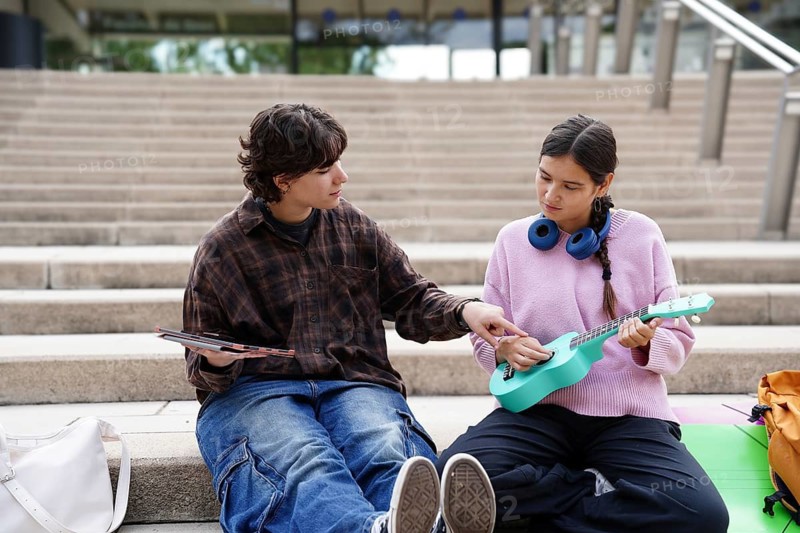 Two teenagers, one non-binary and one female, are situated outside on steps, clearly involved in a creative session with musical and artistic elements. The non-binary teen holds a tablet, while the female plays a ukulele