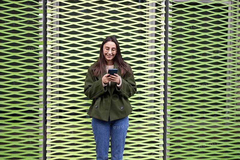 A woman with glasses and a green jacket stands smiling, using her phone against a unique green patterned wall She appears relaxed and engaged with her device