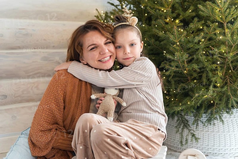 A joyful mother and her young daughter cuddling near a beautifully decorated Christmas tree. Both are smiling at camera