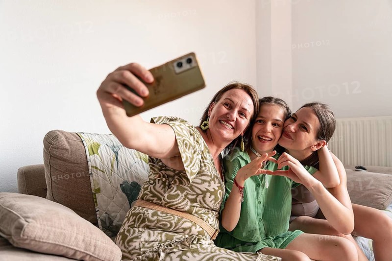 A warm family moment capturing a mother and two daughters, including one with a tracheostomy, taking a selfie while enjoying their time together on a cozy couch