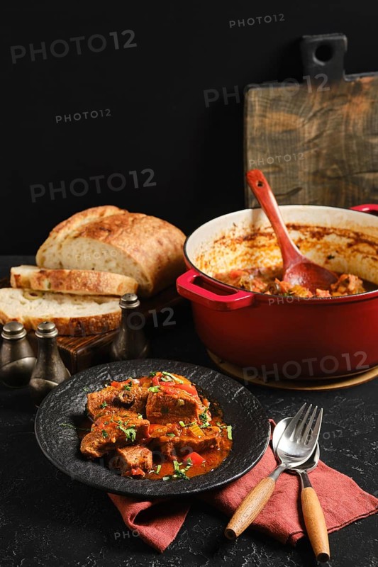 A bowl of beef stew is placed on a black plate next to fresh bread. A pot of stew sits on the table with a wooden spoon. The setting feels inviting and casual