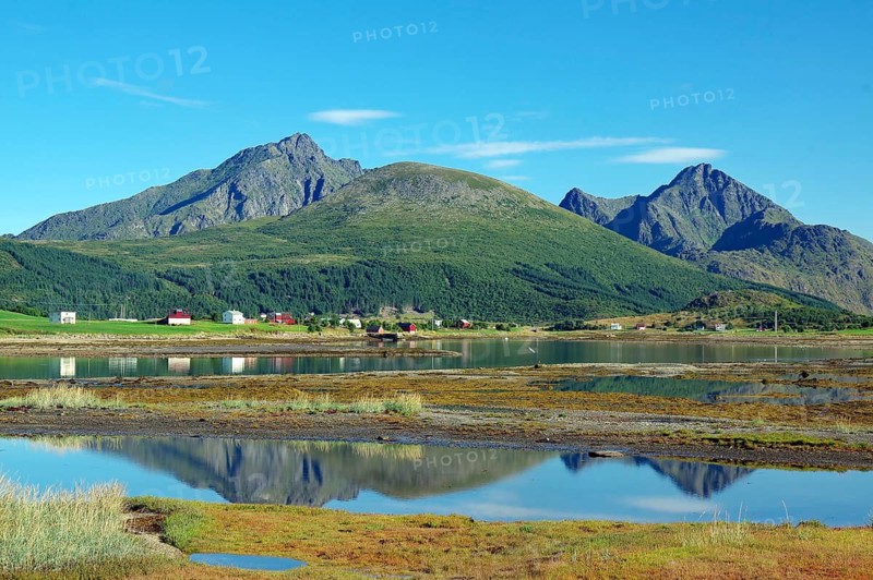A calm body of water reflects mountains and a small village with clear skies, Leknes, Vestvågøya, Lofoten, Nordland, Norway
