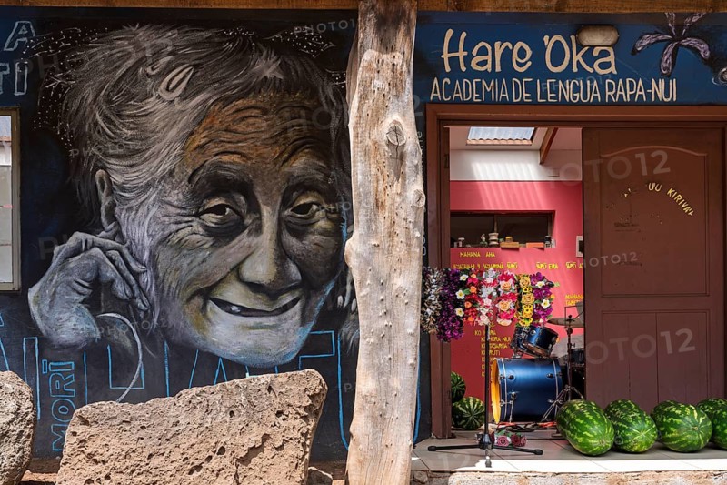 Wall painting with portrait of an elderly smiling woman on the façade of the language school Hare 'Oka — Academia de Lengua Rapa-Nui, with watermelons, drums and flower wreaths in the entrance area, Hanga Roa, Easter Island, Chile