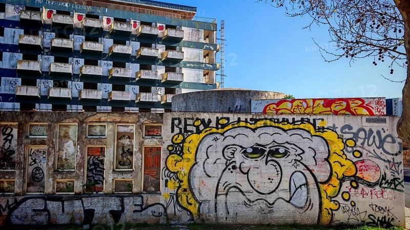 Wall with graffiti in front of urban building, hotel ruin, lost place in the background, blue sky and a tree, Lagos, Algarve, Portugal