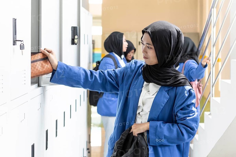 Asian Muslim student in a school hallway, wearing a blue uniform, organizing her belongings in a locker. She is preparing for a day of learning with a focused expression